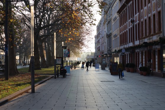 Die Dresdner Hauptstraße. Links sind Laubbäume mit orange-goldenen Blättern. In der Mitte mehrere Silhouetten von Menschen, die sich im Gegenlicht auf der mit Gewehgplatten ausgelegten Fußgängerzone fortbewegen. Rechts Fassaden in Rot- und Orangetönen.