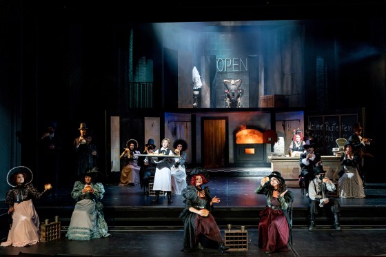 Gesamtansicht des dunkel gehaltenen Bühnenbildes. Im Vordergrund verteilt sitzende Mitglieder des Chores in Gothic-Kleidung. Weiter hinten der Bühnenaufbau: Im Erdgeschoss eine Theke, an der Mrs. Lovett mit roten Haaren steht. Dahinter der glühende Ofen. Im Obergeschoss der Barbiersalon. Sweeney Todd sitzt im Barbier-Stuhl.