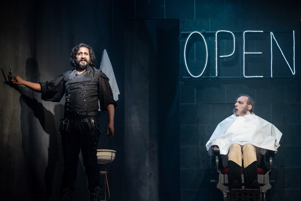 Aufnahme aus größerer Nähe im Obergeschoss des Bühnenbaus, im Barbiersalon. Links steht Sweeney Todd und hält die Klinge auf Schulterhöhe von sich weg und singt. Rechts sitzt Richter Turpin mit umgelegten weißen Tuch bereit zur Rasur und blickt zu Sweeney Todd. Dahinter eine schwarz geflieste Wand und weiß leuchtende Buchstaben des "Open"-Schriftzuges.